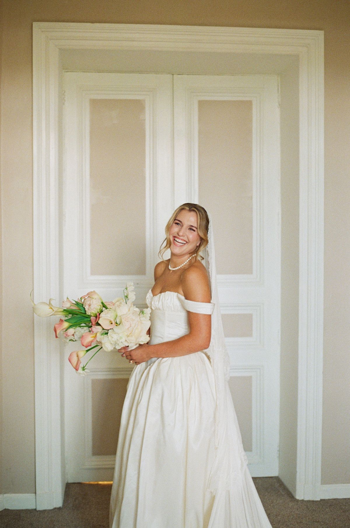 The bride in an off-the-shoulder white gown holds a pale bouquet and smiles in front of tall white double doors.