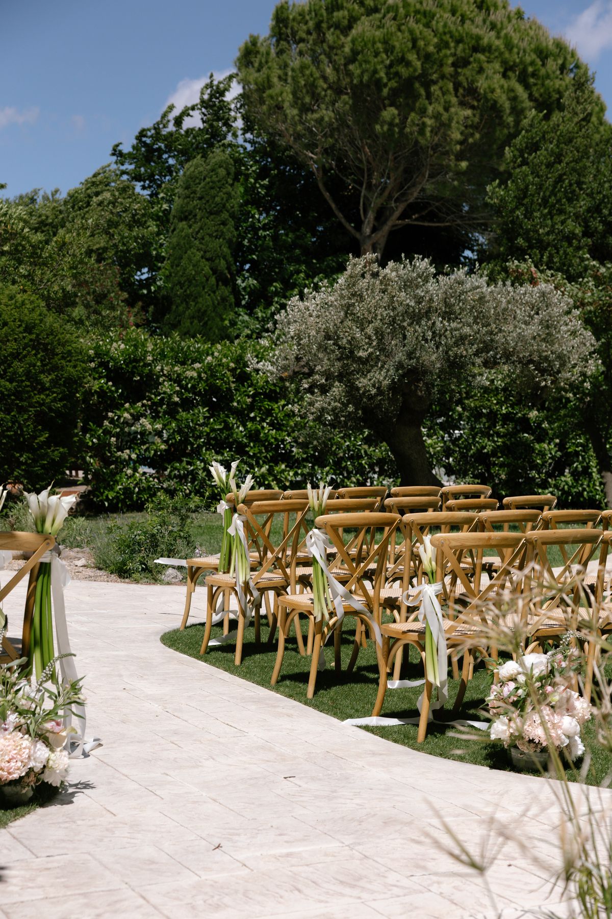 Empty wooden cross-back chairs with white calla lilies and ribbons line a stone aisle on a lawn, set for an outdoor ceremony.