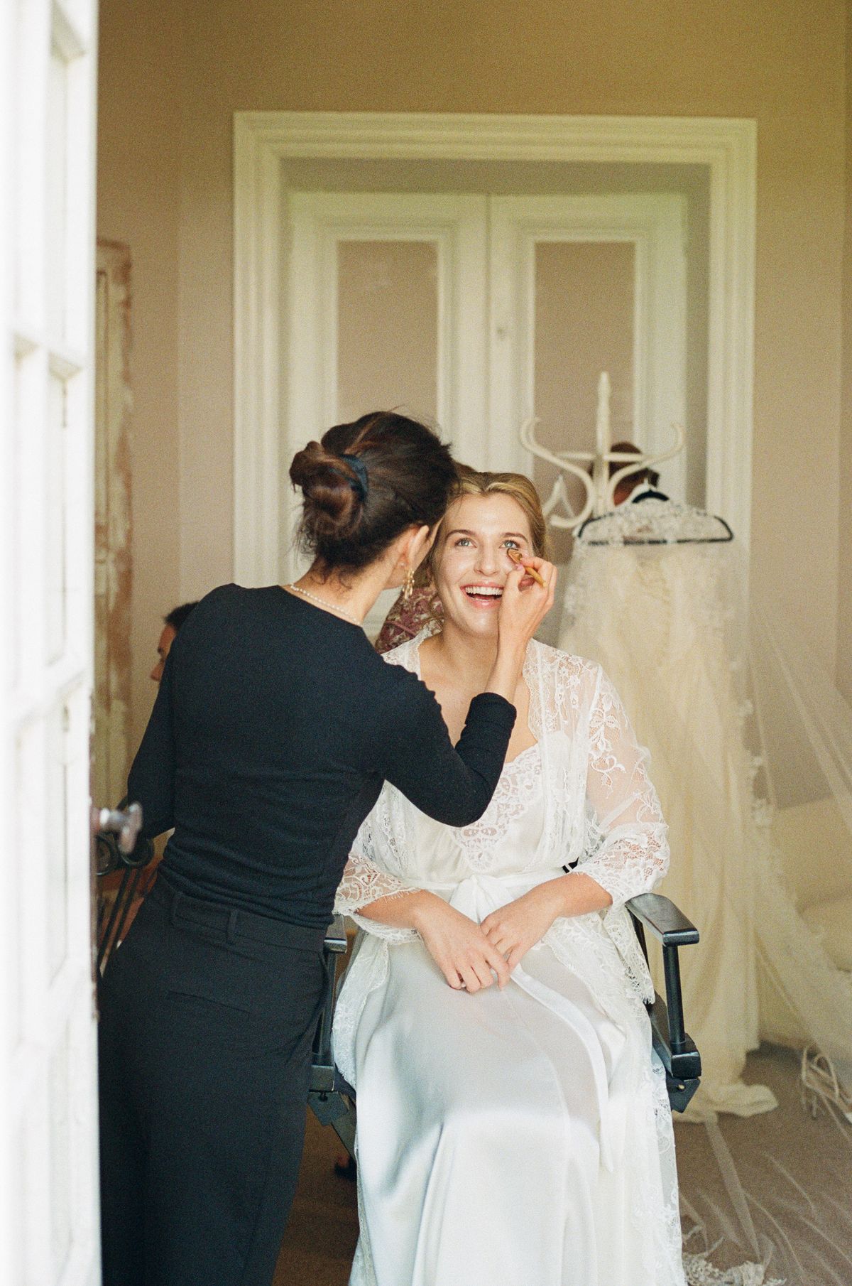 The bride sits laughing in a robe while a make-up artist dressed in black attends to her face beside a tall doorway.