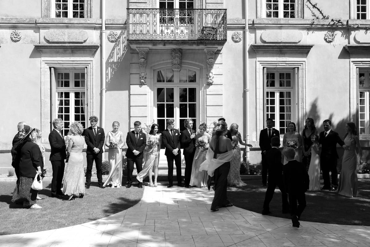 A large group of guests in formal clothes stand together on a paved terrace in front of a chateau with a balcony.