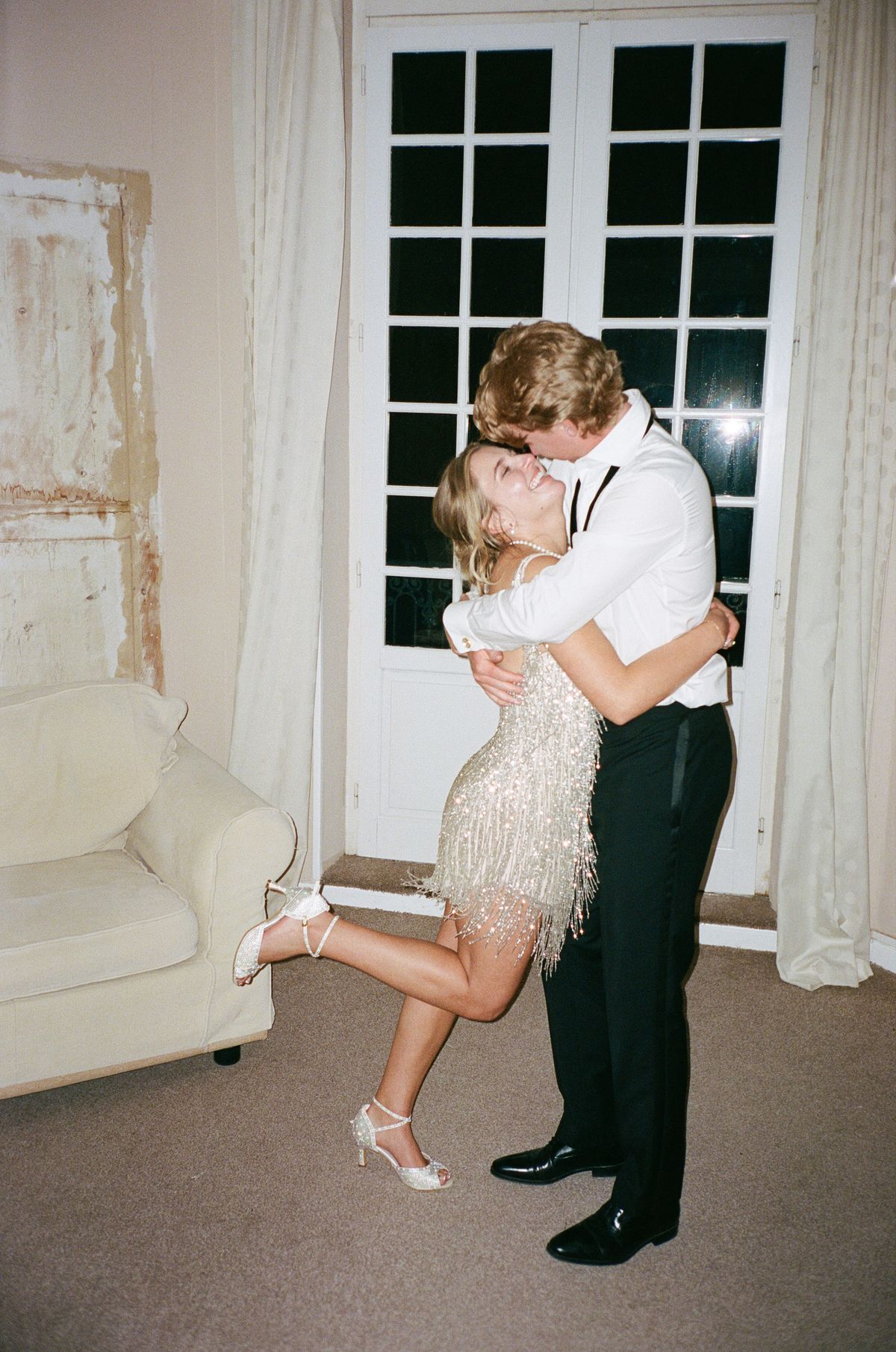 The groom in a white shirt kisses the bride in a short fringed dress, who lifts one heel, near French windows.