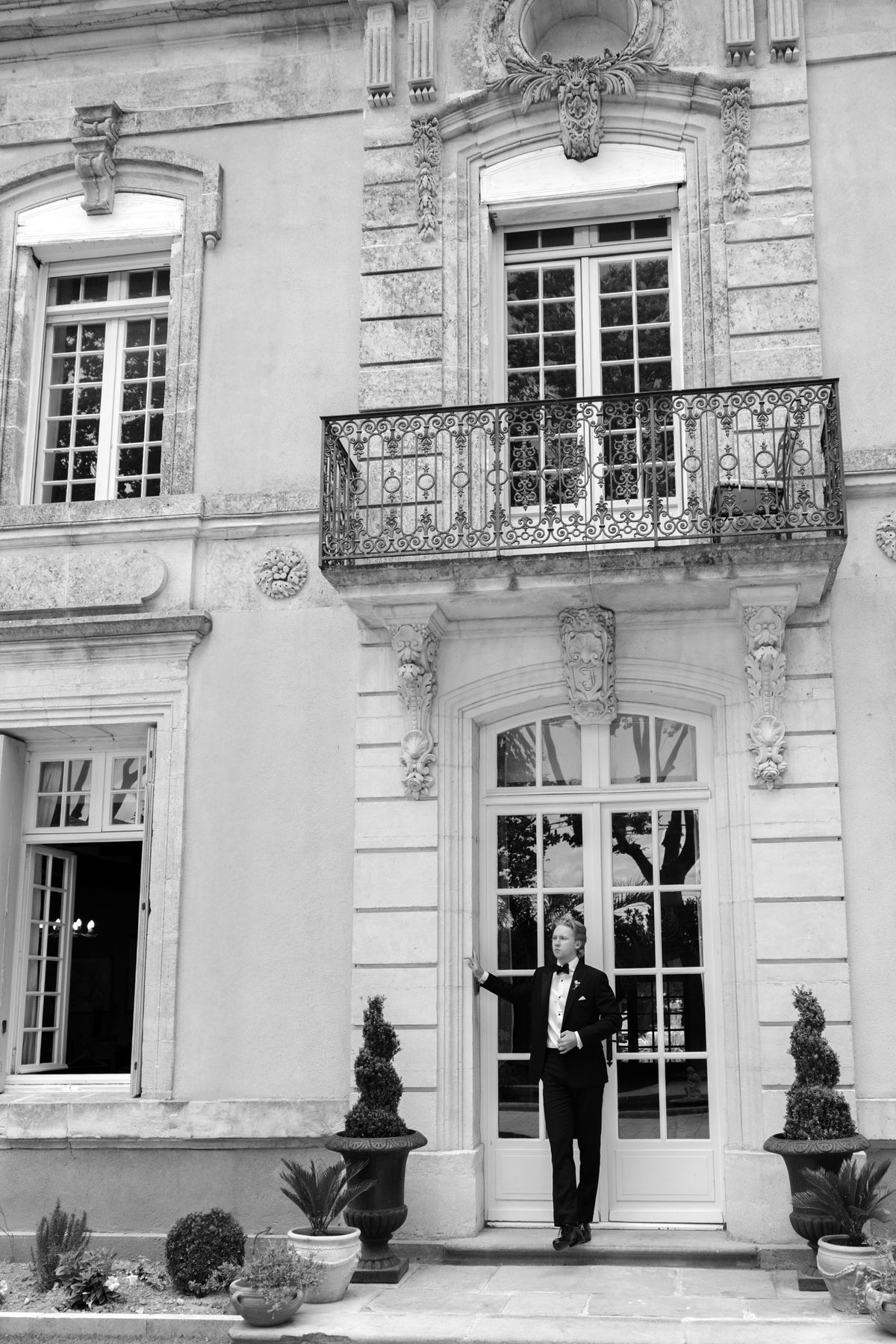 In black and white, the groom stands in a doorway beneath an ornate balcony on a stone chateau, flanked by potted topiary.