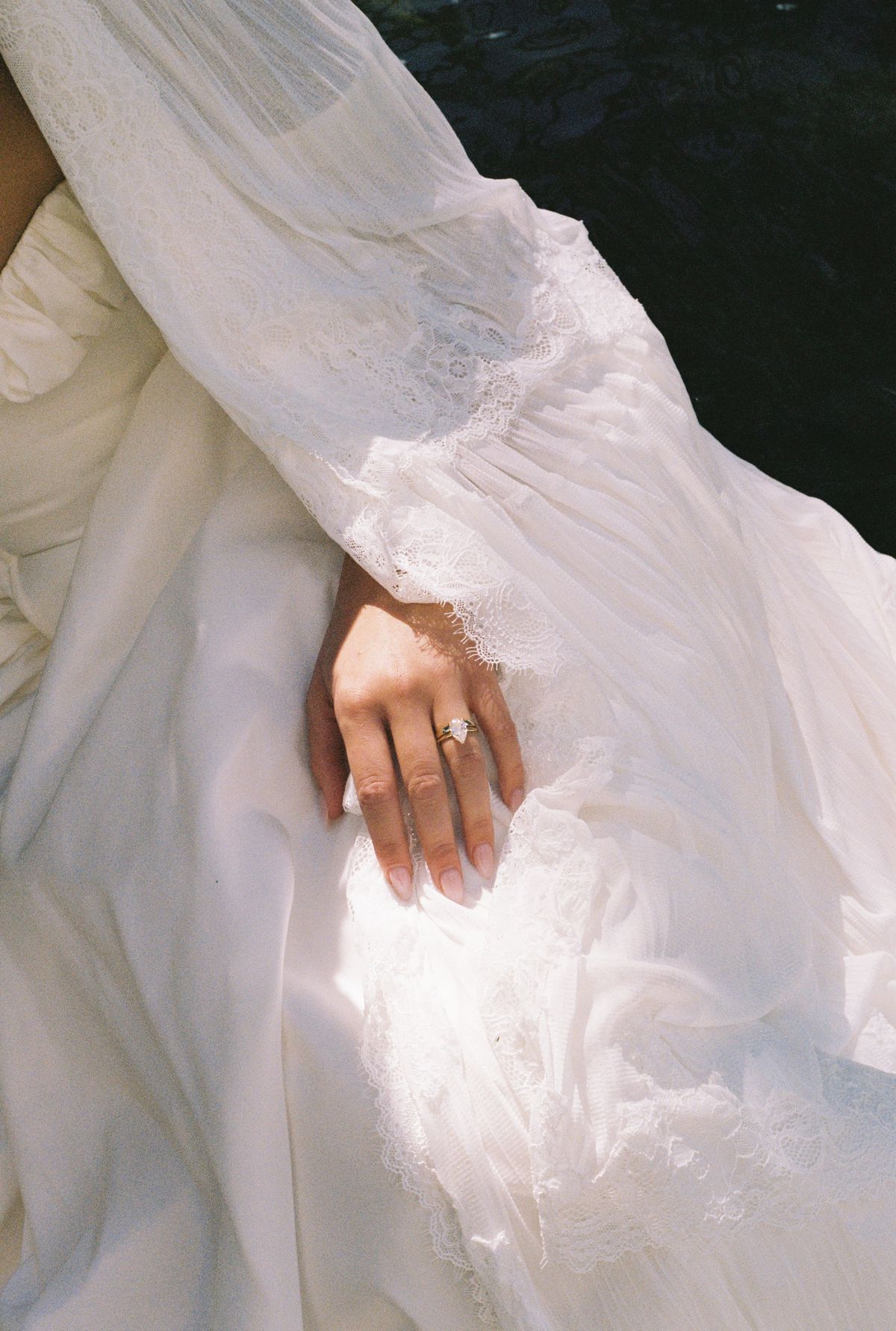 The bride's hand, wearing a ring, rests on her white dress, with a sheer lace-trimmed sleeve draped over her arm.