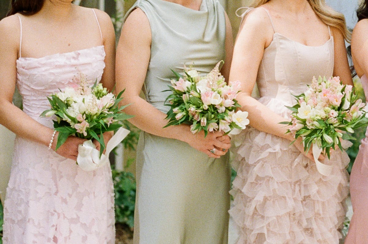 Three bridesmaids, shown from the shoulders down, wear pink and sage green dresses and hold bouquets of pale pink flowers.