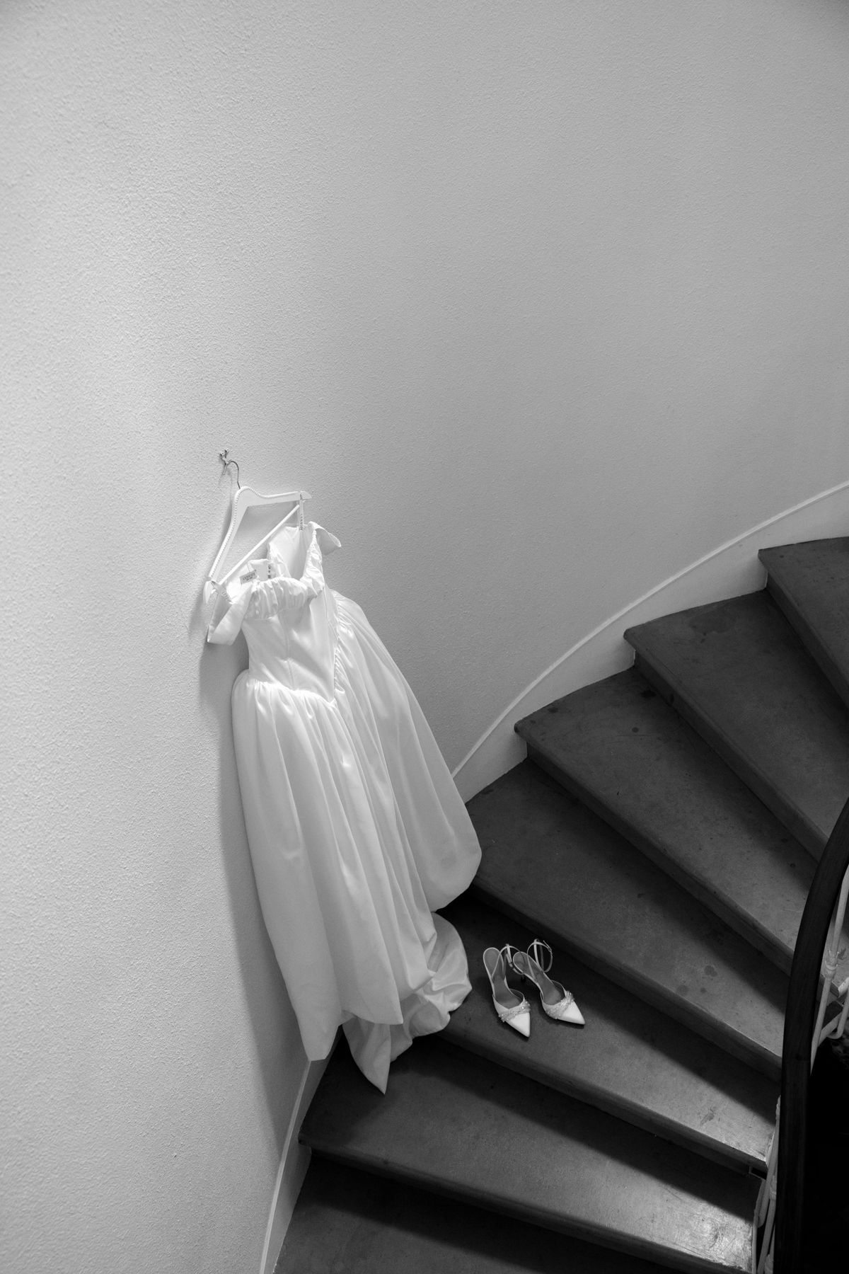 In black and white, a wedding dress hangs on a wall beside a dark staircase, with a pair of shoes on the steps.