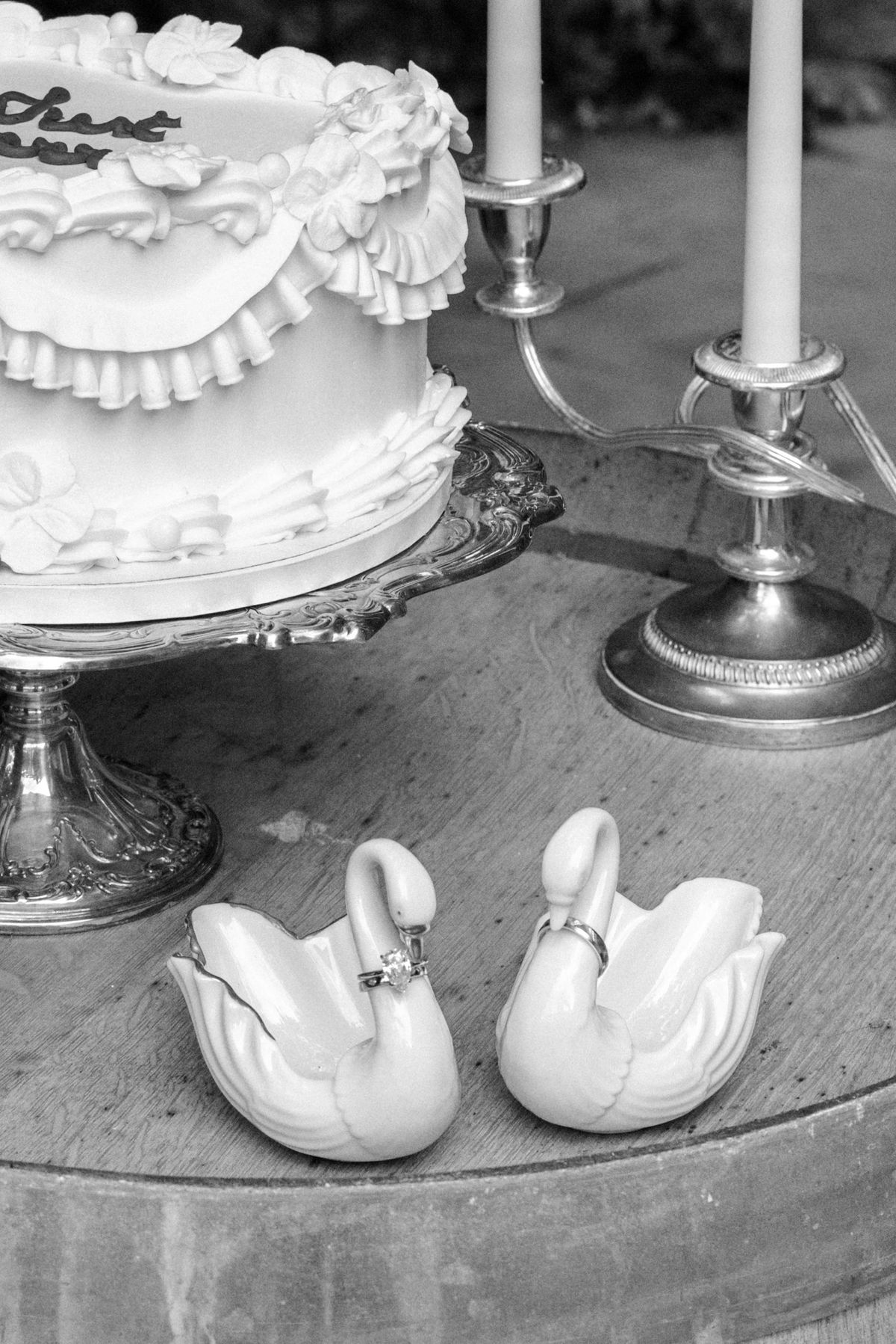 A small frilled cake stands beside two tall candlesticks and a pair of white swan figurines wearing rings, on a wooden barrel top.