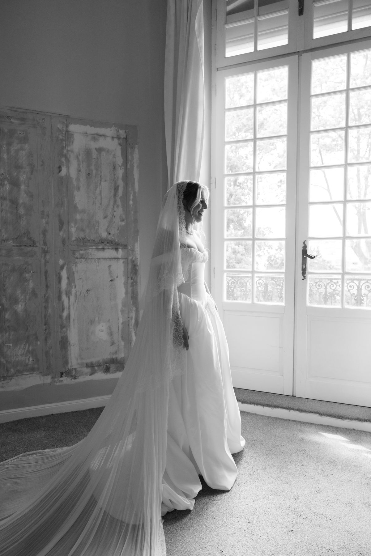 In black and white, the bride in a long veil stands beside a tall French window in a bright room.