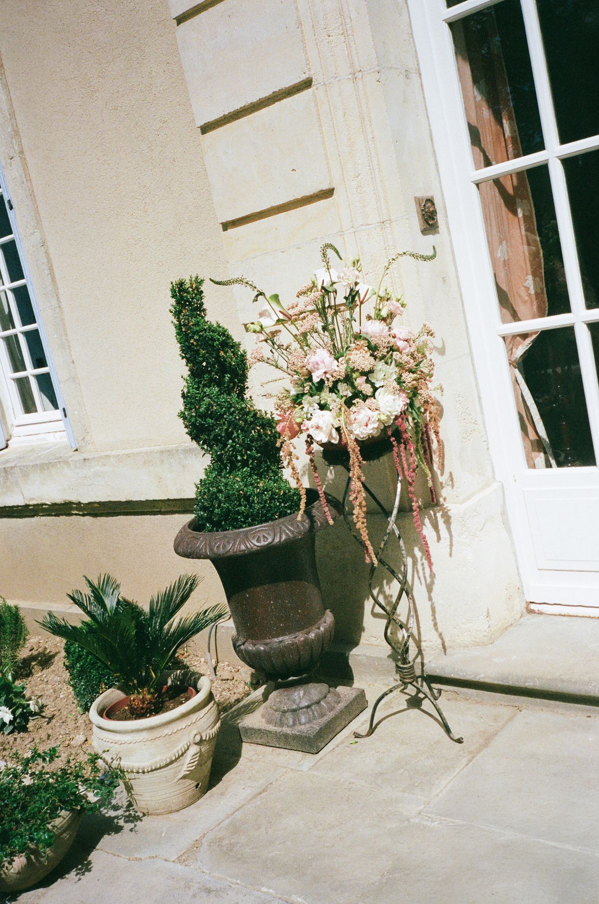 A large arrangement of pink and white flowers on a tall stand beside a clipped topiary in a stone urn near a doorway.