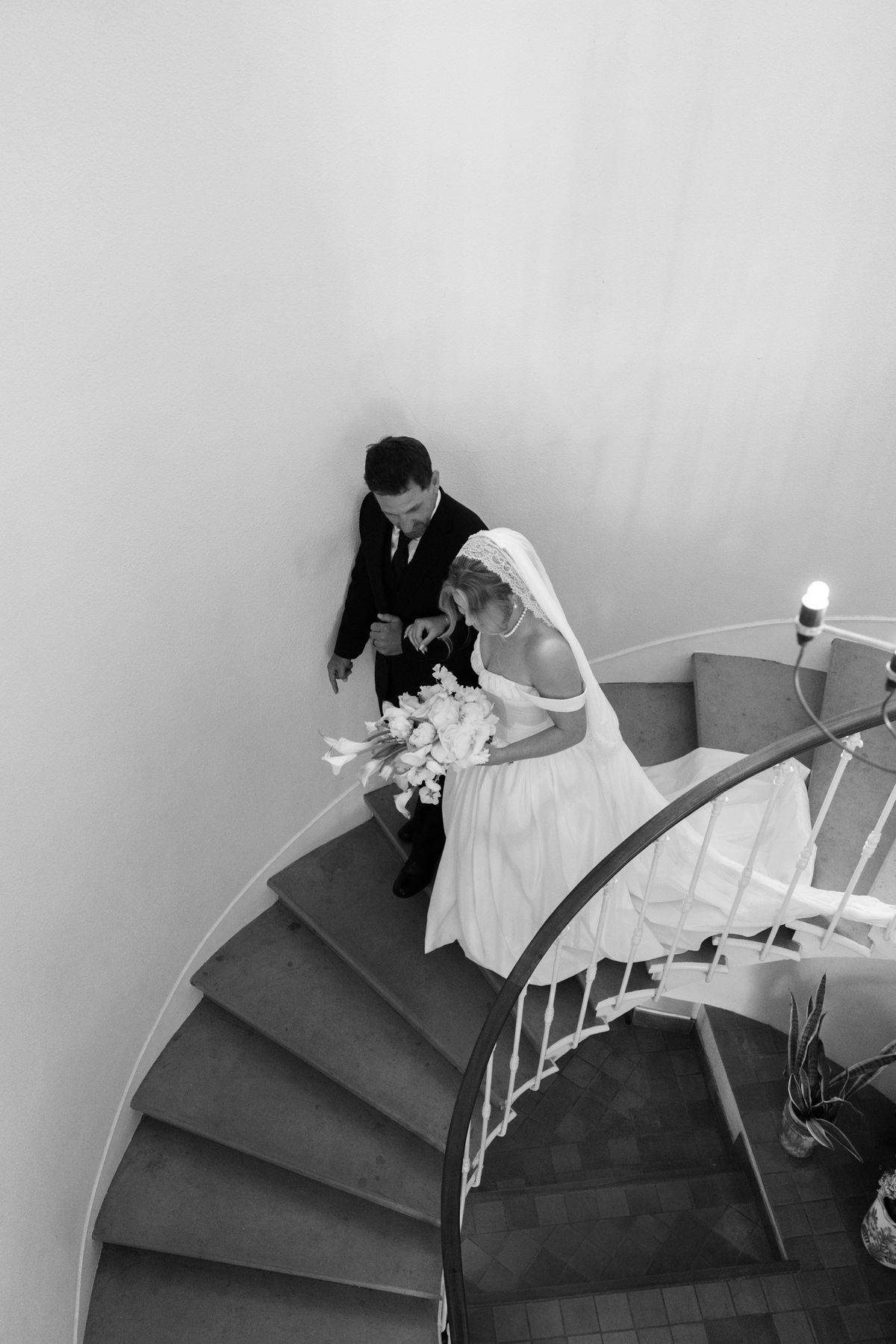 In black and white, seen from above, the groom leads the bride holding a bouquet down a curving staircase with white walls.