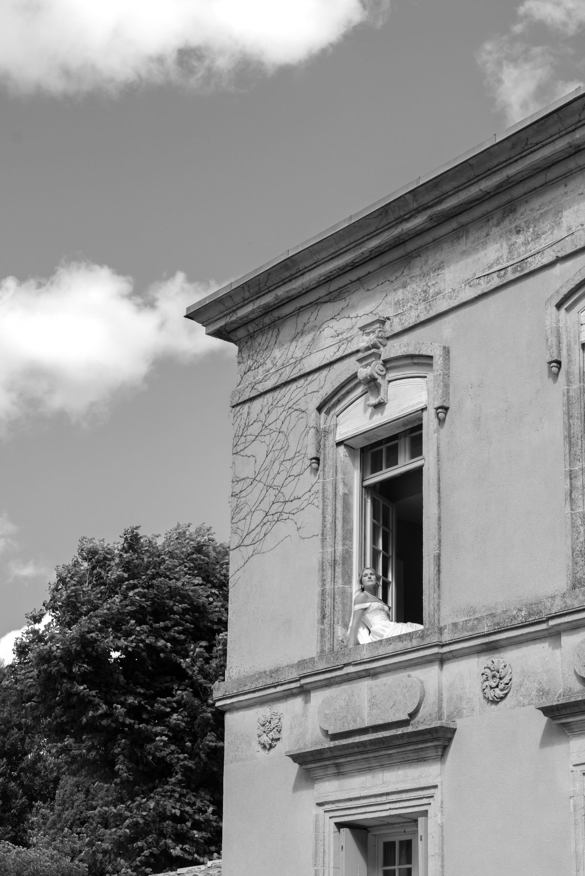 A woman in a light gown sits at an open upper window of a stone building, beside a leafy tree, in black and white.