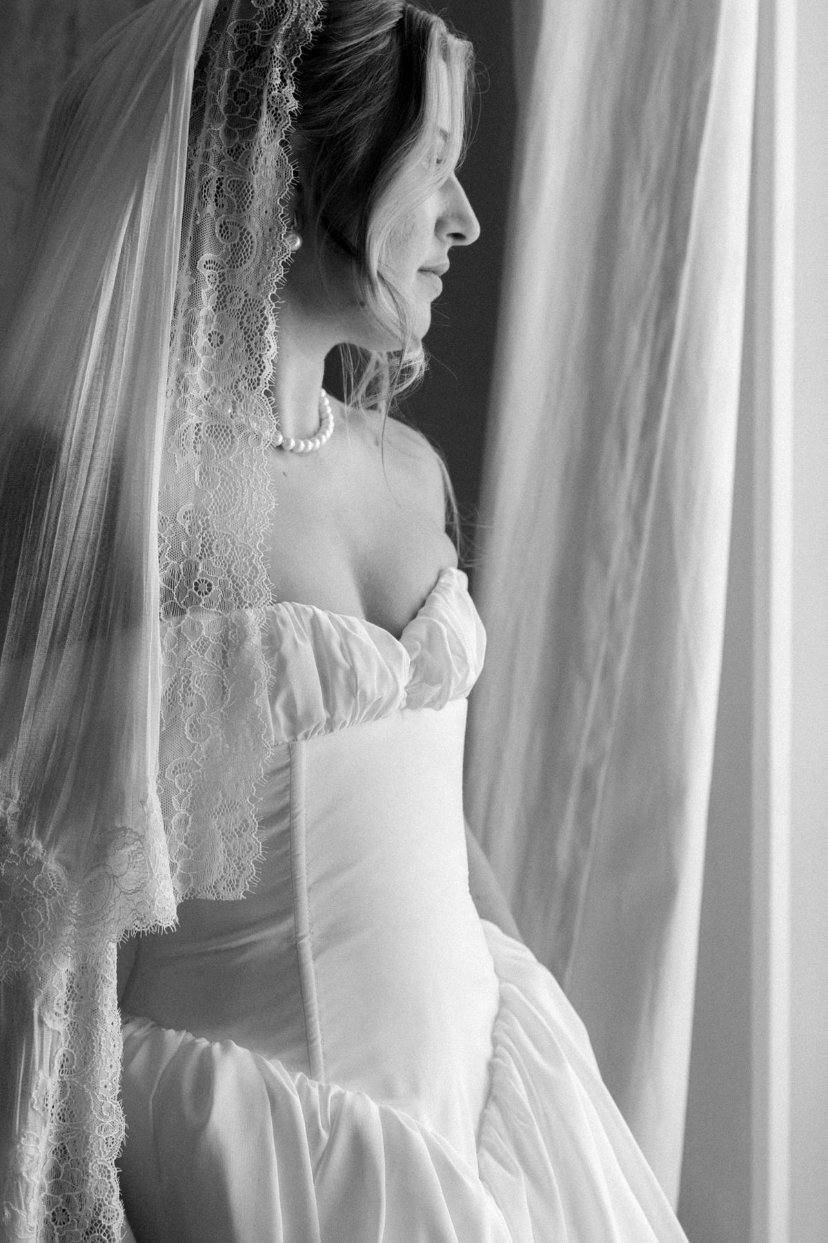 The bride in profile, wearing a lace-edged veil, pearl necklace and strapless gown, gazing towards a softly draped curtain.