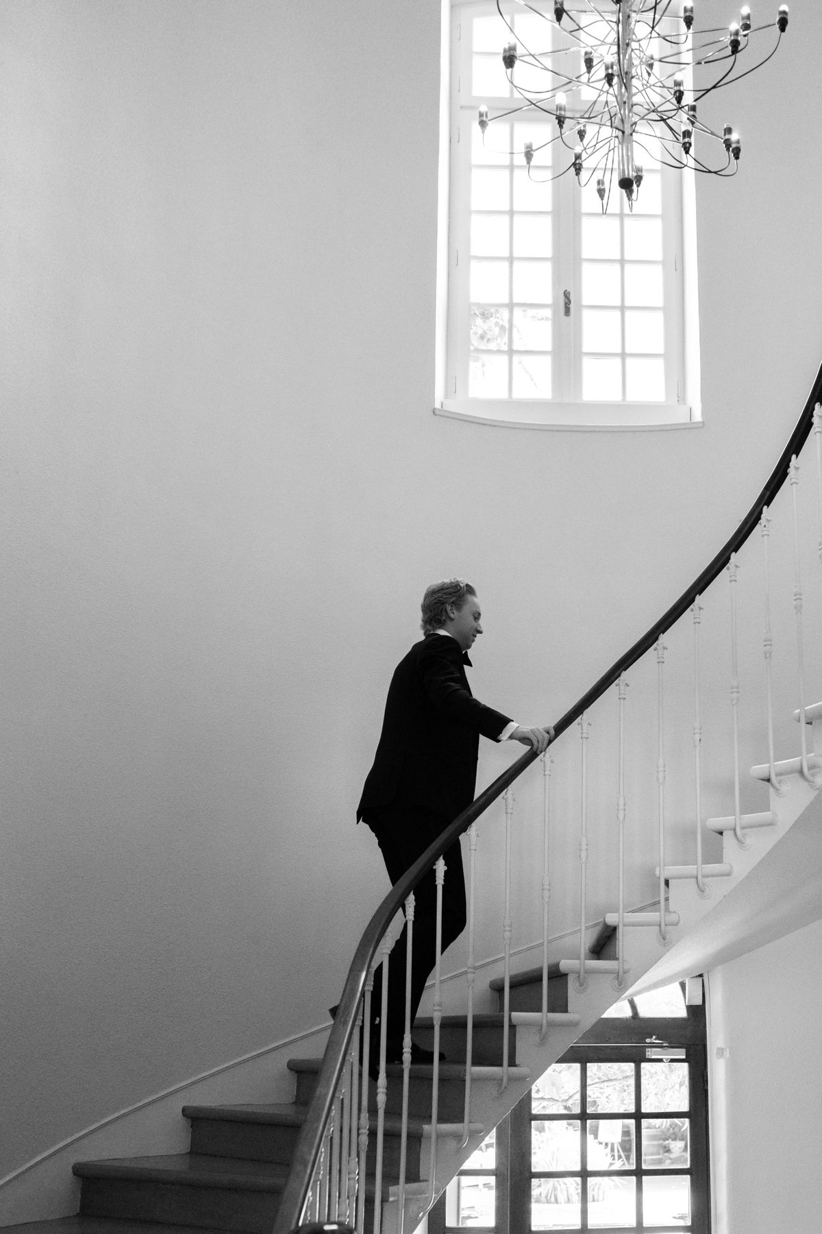 A man in a dark suit and bow tie climbs a curved staircase, hand on the rail, beneath a tall window and chandelier.