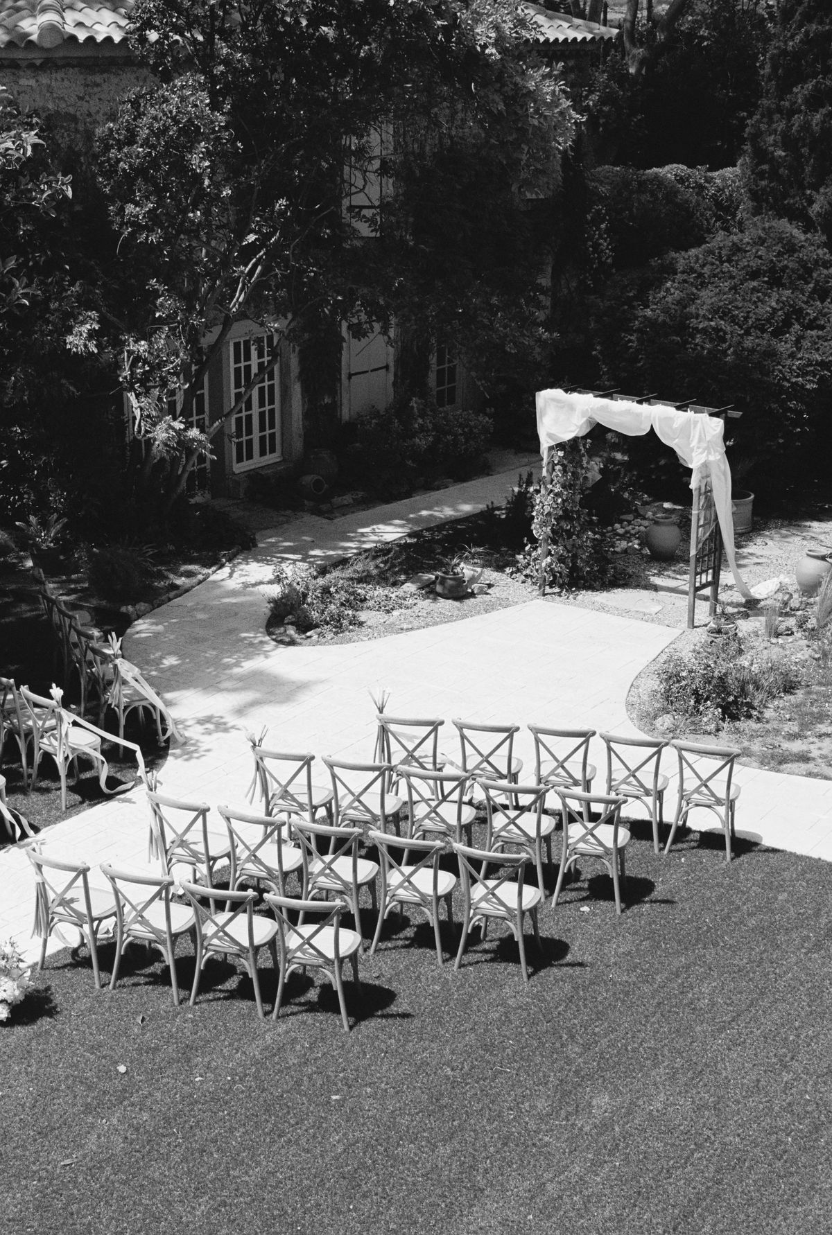 A high view shows rows of cross-back chairs on a lawn beside a white path leading to a floral arch near a stone building.