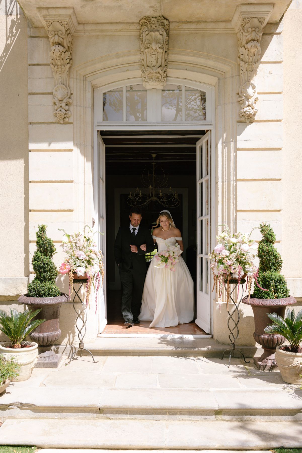 The bride in a white gown and a man in a dark suit stand in an ornate stone doorway framed by flowers and topiary.