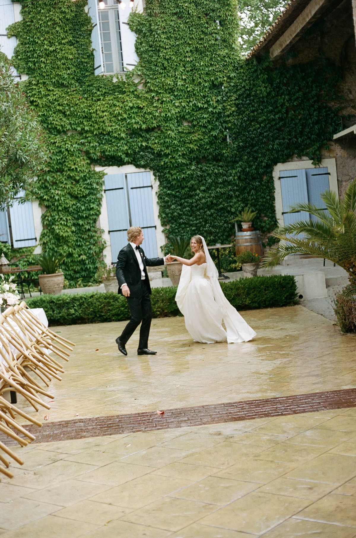 The groom holds the bride's hand as she twirls in her gown on a courtyard paved in stone, with ivy-clad walls behind.