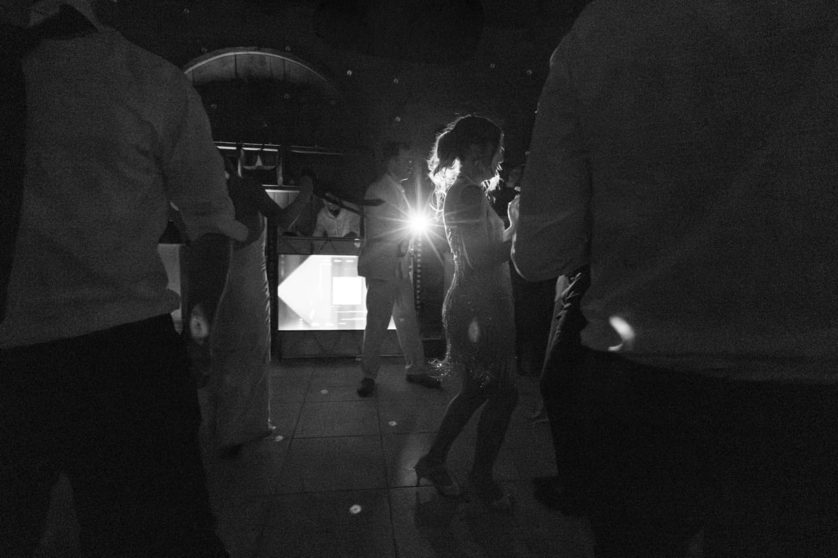 Silhouetted guests and the bride dance in a dim room, with a bright burst of light behind her.