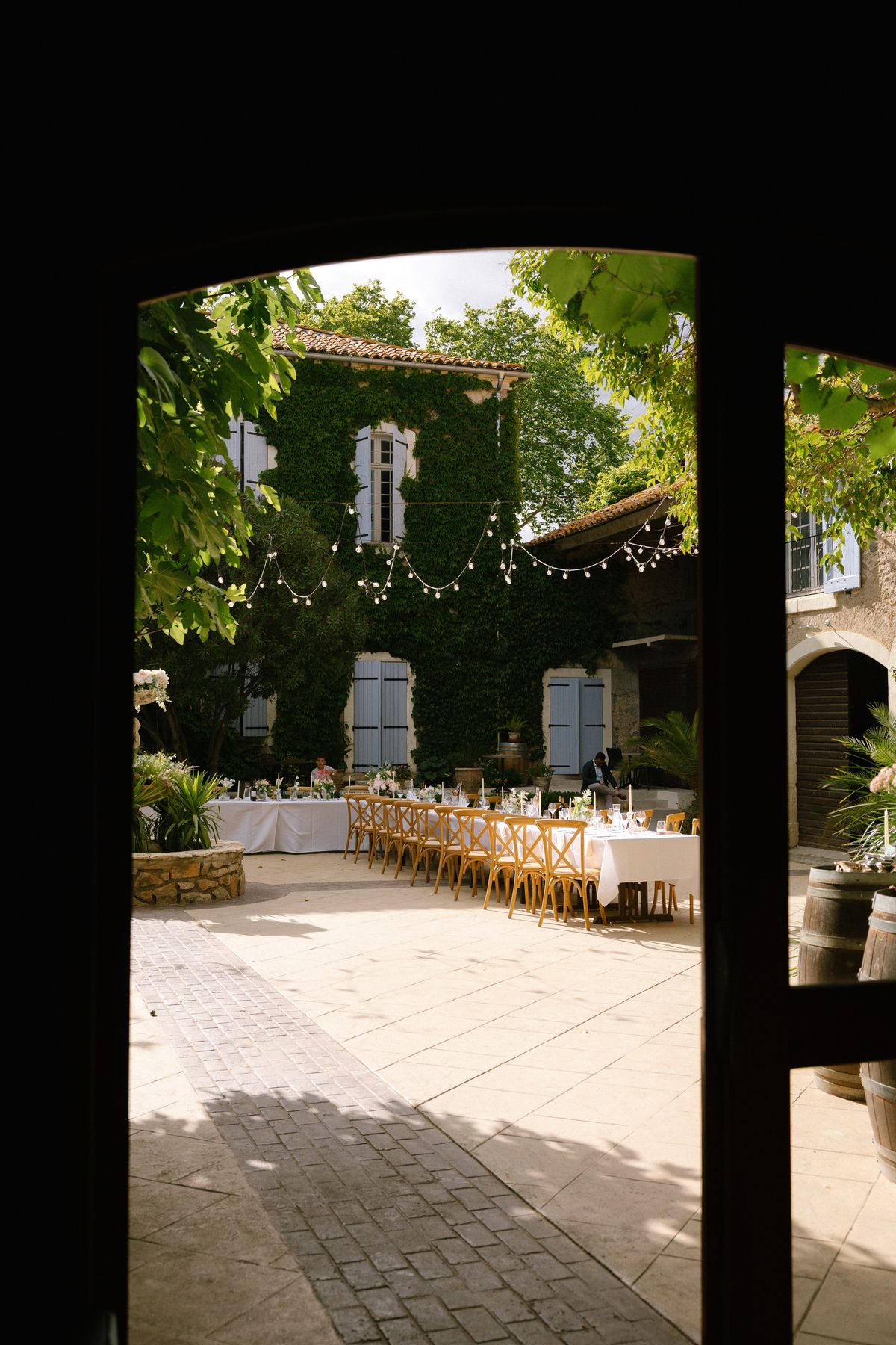 A view through an open doorway onto a courtyard set with long tables, wooden chairs and string lights.