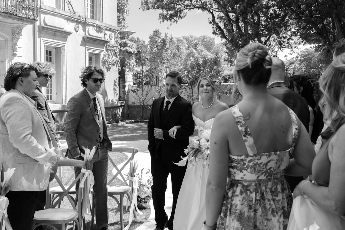 The bride and groom stand among smiling guests next to wooden chairs with ribbons, under trees near a stone building.