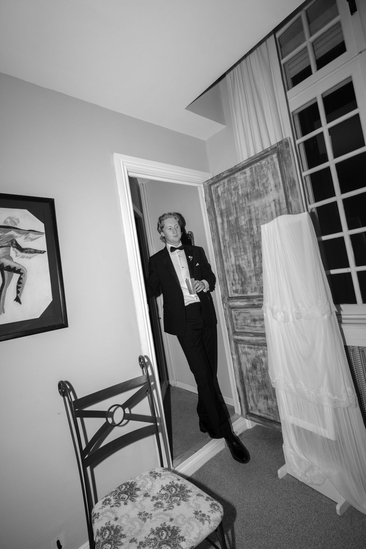 The groom leans against a doorway holding a glass, next to a chair and a white dress hanging nearby.