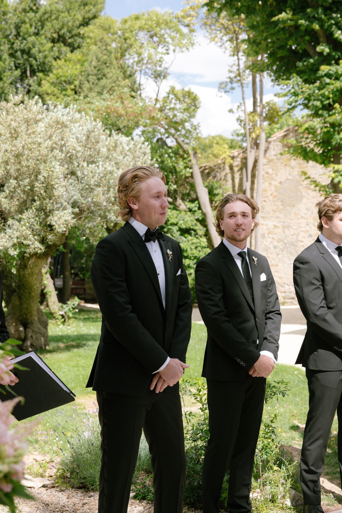 Three men in dark suits and bow ties stand in a sunlit garden, looking ahead, while an officiant holds a book at the edge.