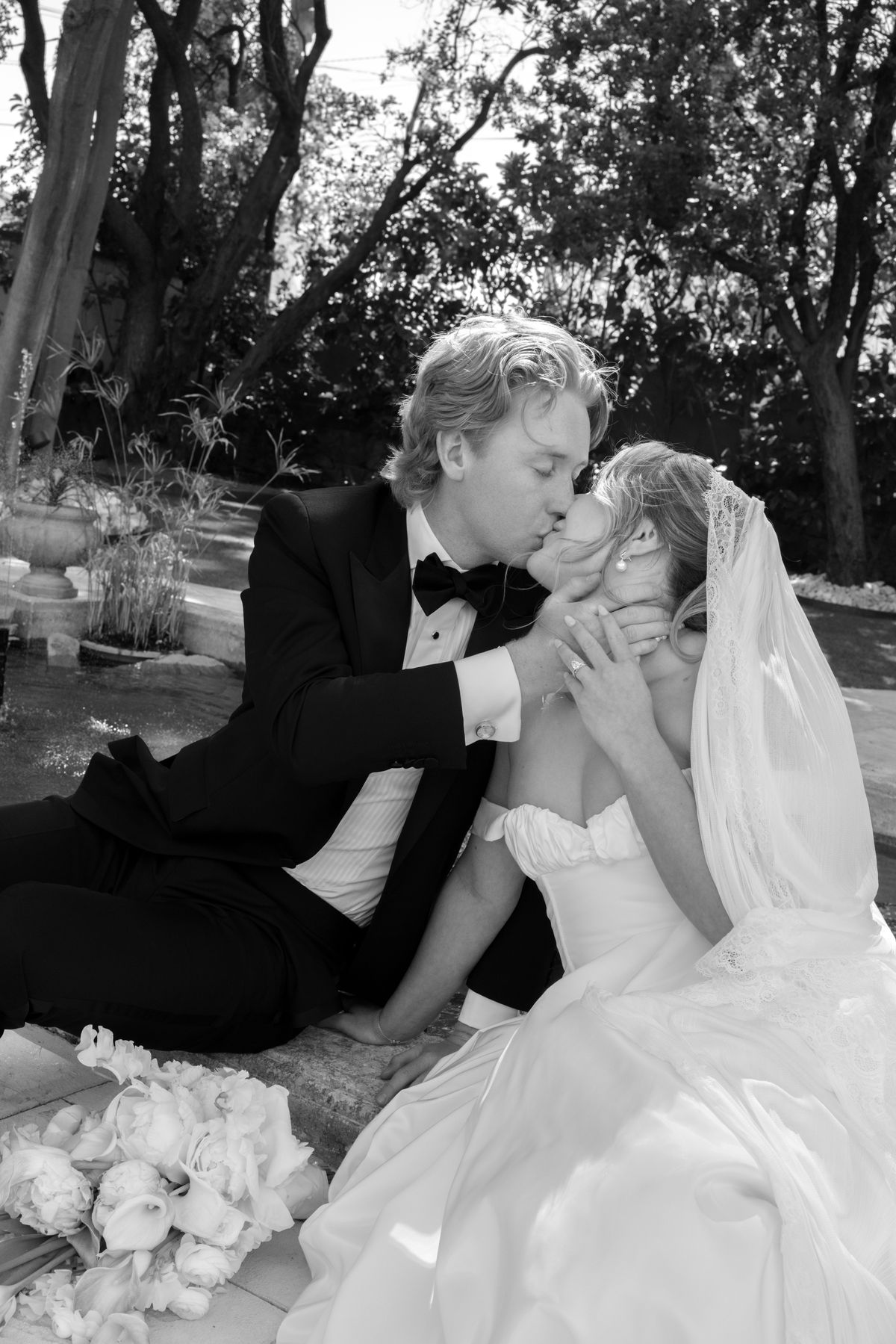 The groom in a tuxedo kisses the bride, who wears a veil, as they sit on the ground beside bouquets under trees.