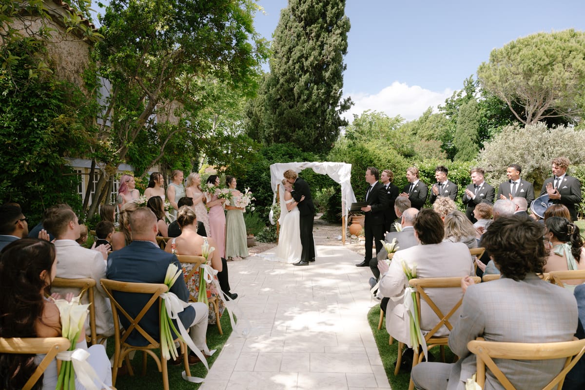 Guests sit on wooden chairs either side of a sunlit garden aisle as the couple stand beneath an arch at the far end.