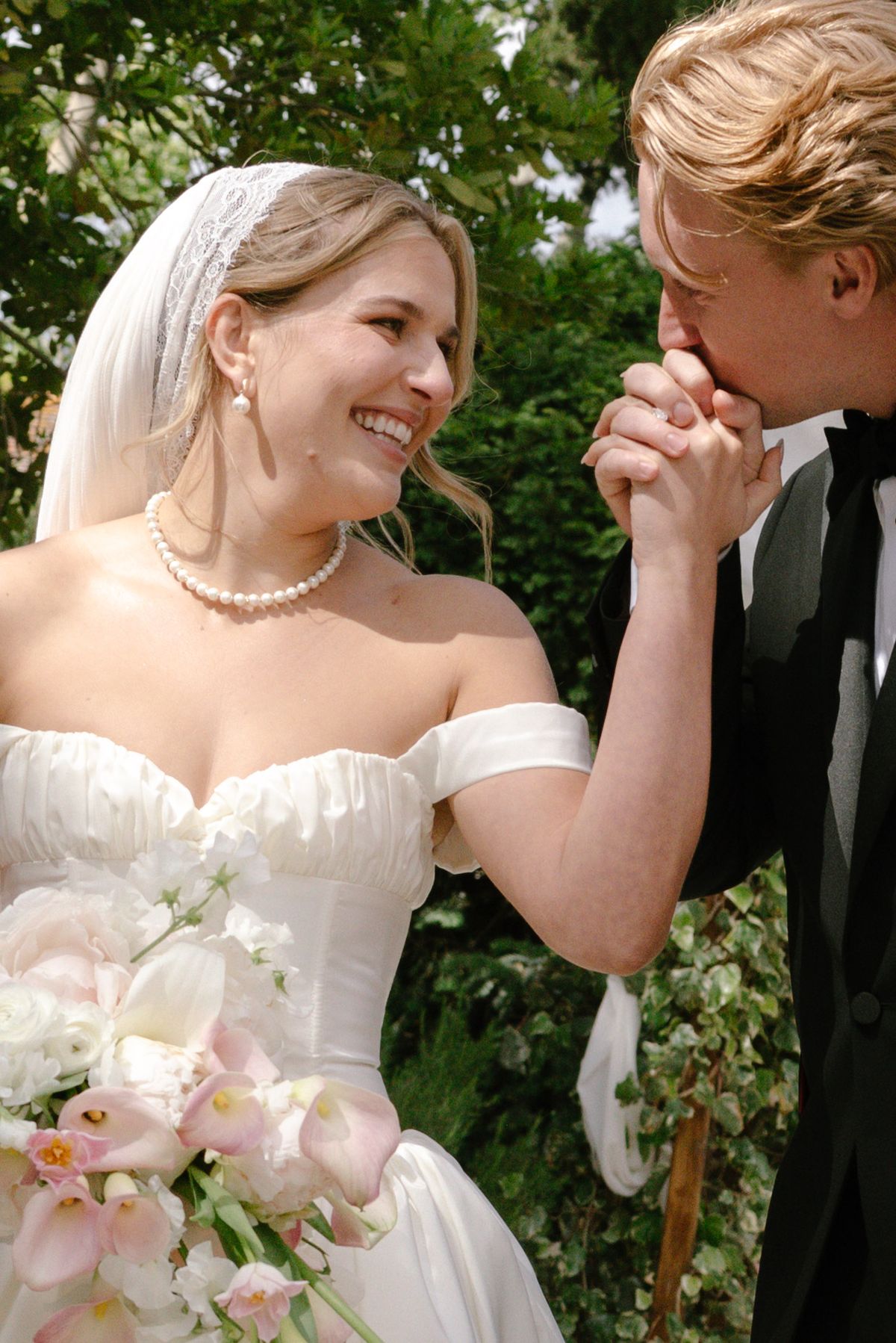 The smiling bride in a pearl necklace and veil holds a pink bouquet as the groom kisses her hand.