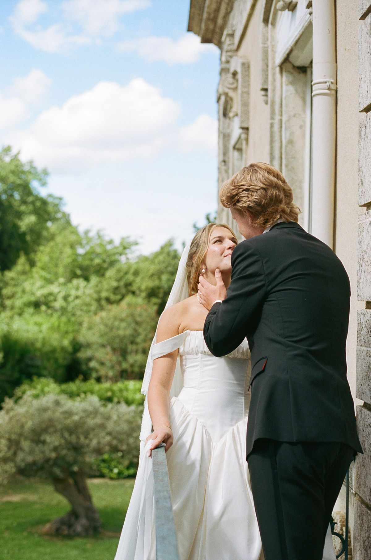 The groom in a black suit leans towards the bride as he adjusts her gown, with greenery and blue sky behind.