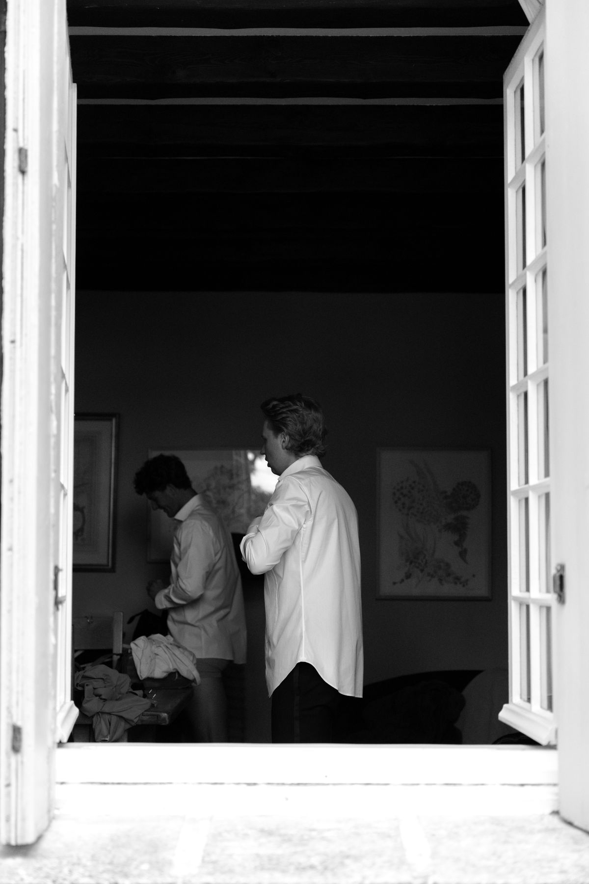 In black and white, seen through open doors, two men in shirtsleeves get dressed in a dimly lit room.