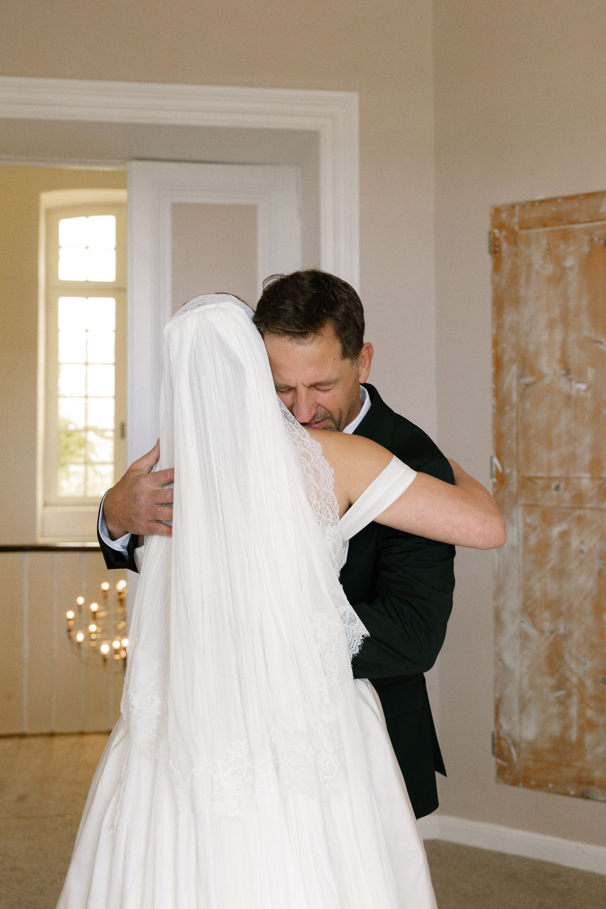 The bride in a long veil hugs a smiling man in a dark suit in a bright room with tall doors.