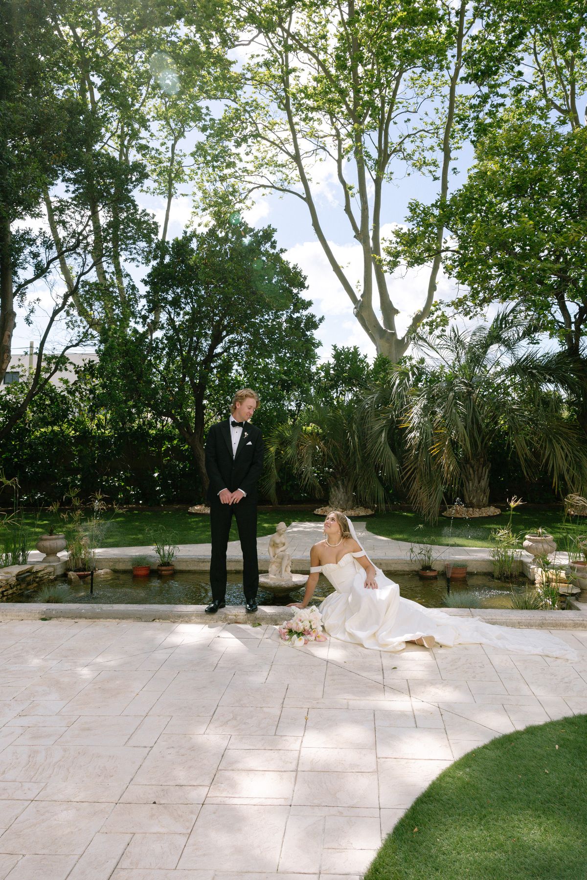 The groom in a dark suit stands on a pale stone terrace while the bride sits on the ground in her gown, trees rising behind them.