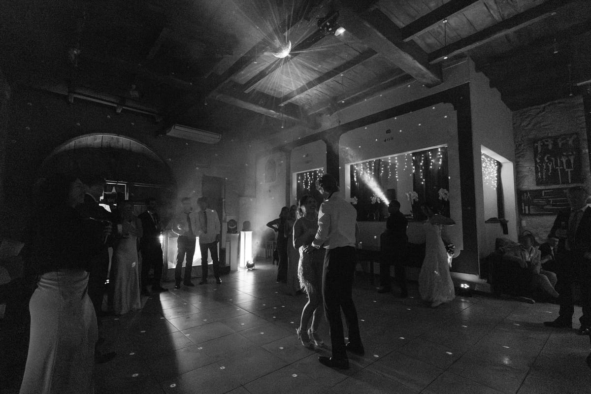 In black and white, guests stand on a dance floor in a dim hall lit by spotlights and string lights.