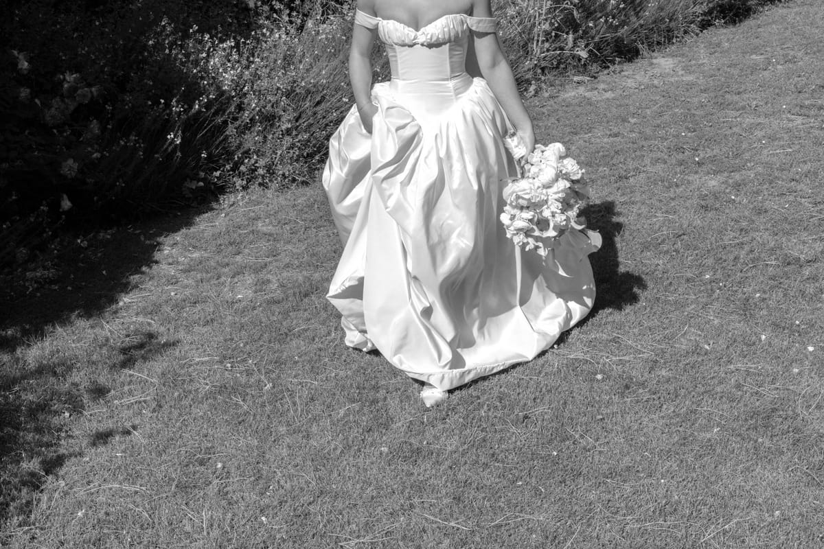 In black and white, the bride's full skirt and bodice are seen from behind as she walks over grass holding a bouquet.