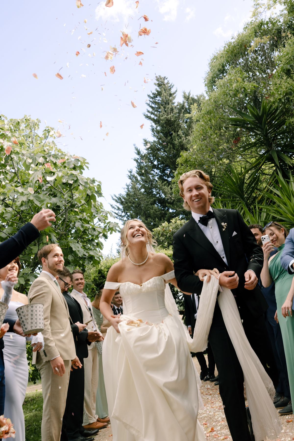 The smiling couple walk through a crowd of guests as pink petals fly through the air, the bride holding a long white sash.
