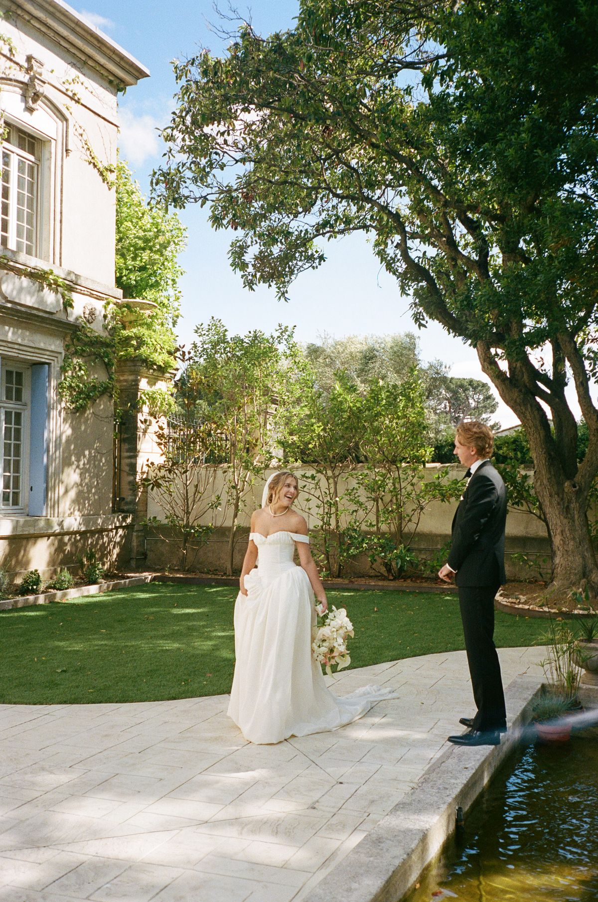 The bride in an off-the-shoulder white gown stands on a stone terrace facing the camera while the groom stands a short distance away on the lawn.