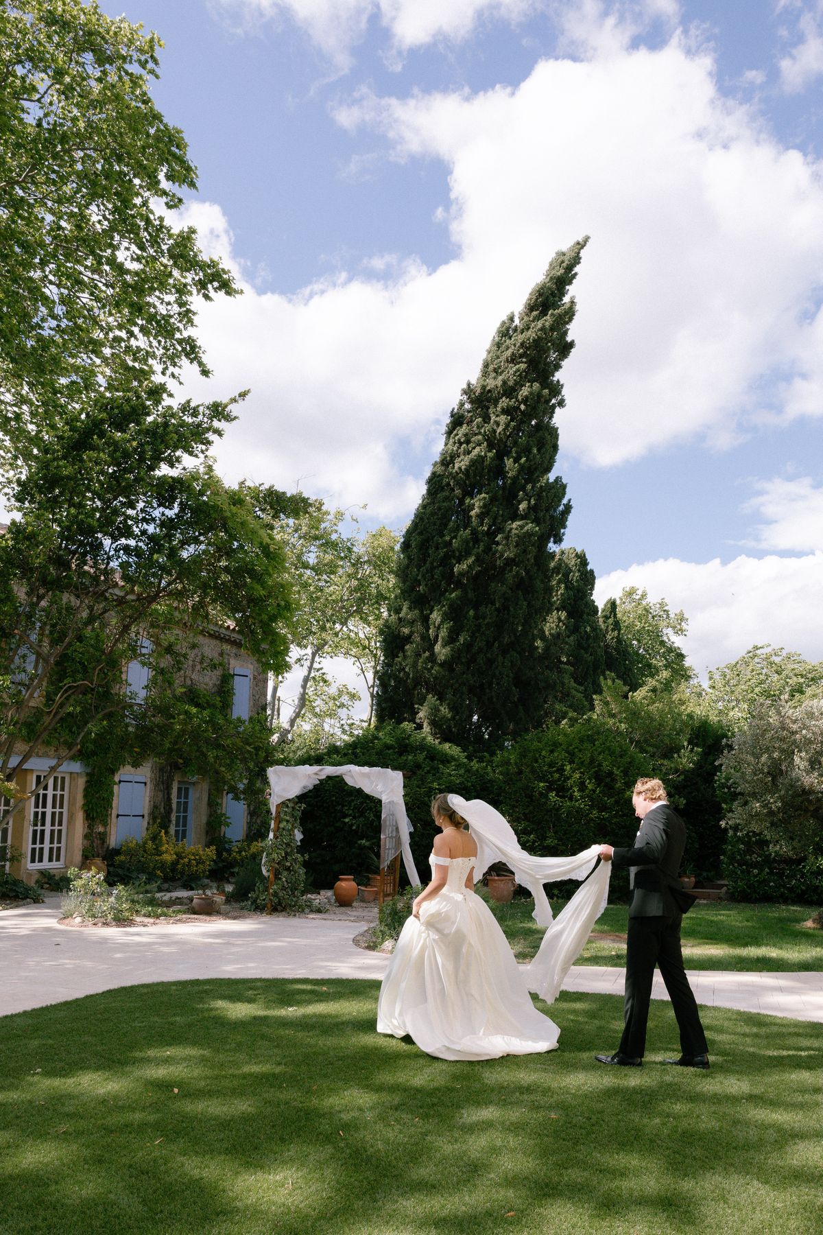 The bride holds her veil out in the breeze on a green lawn as the groom walks beside her, with a chateau and tall cypress behind.