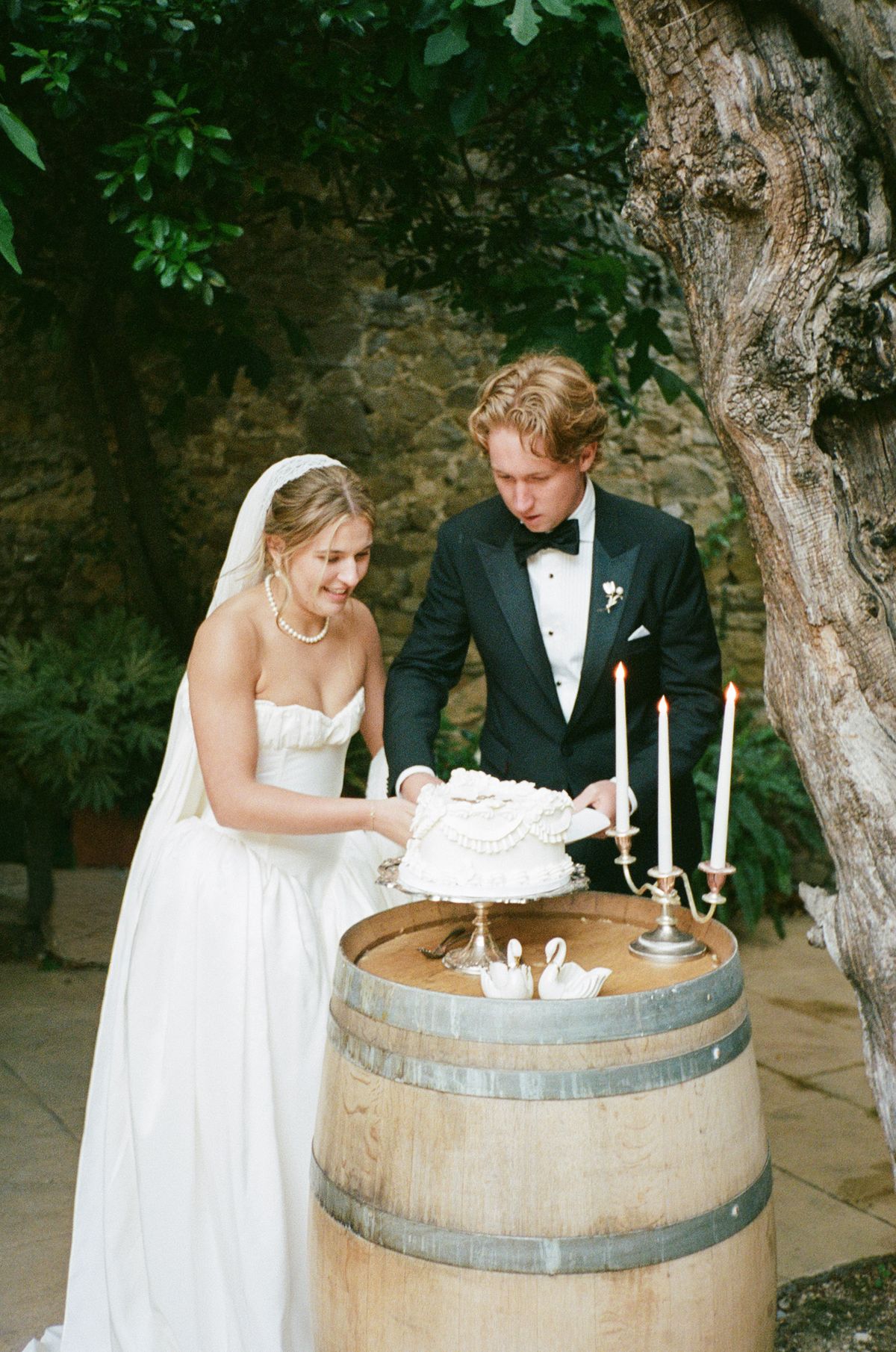 The bride and groom cut a small white cake set on a wooden barrel with candles, beneath trees.