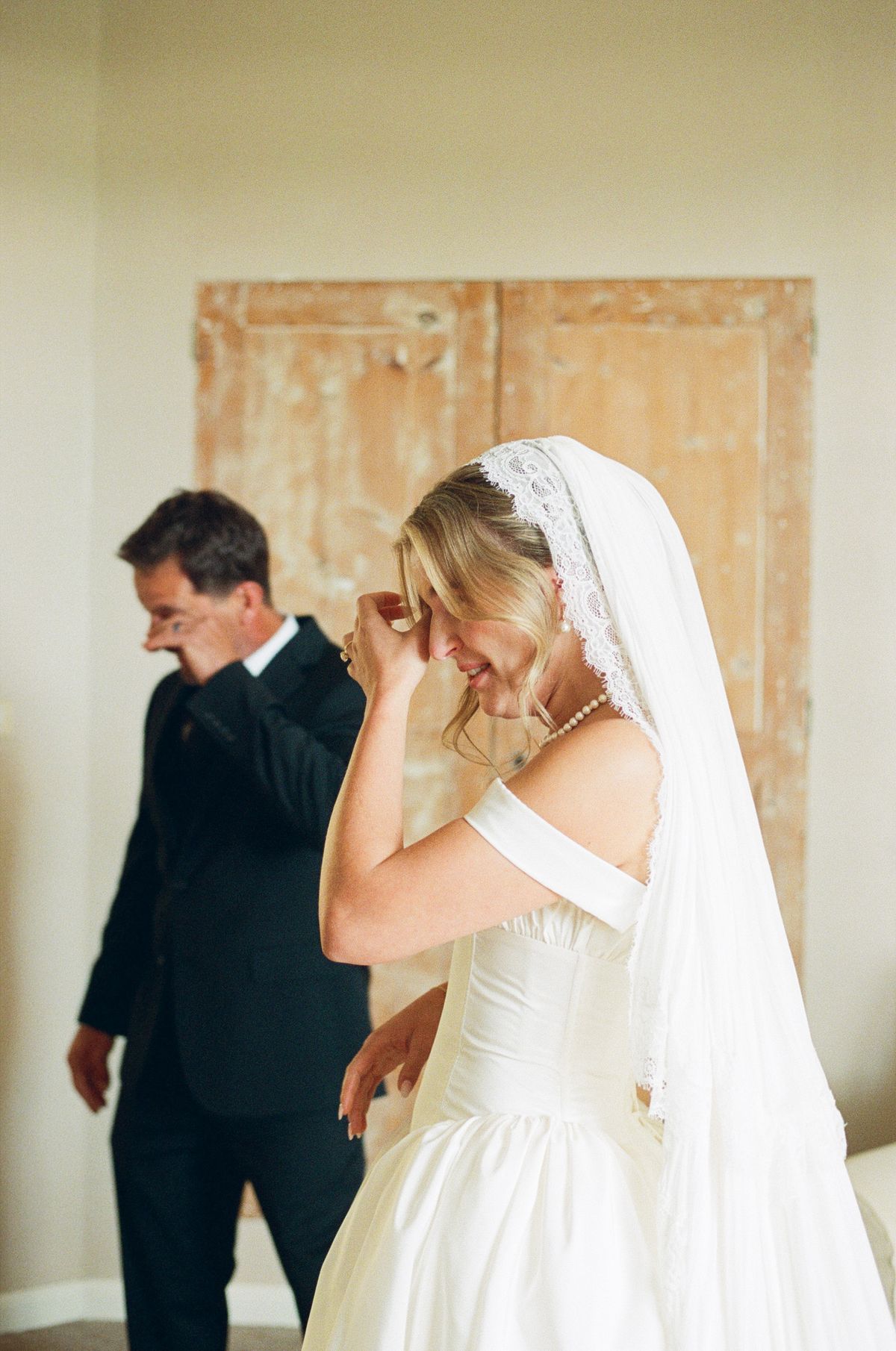 The bride in a veil raises a hand to her face, visibly moved, while a man in a dark suit stands beside her.