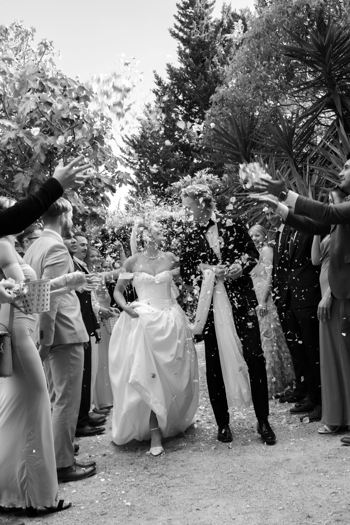 In black and white, the couple walk between two lines of guests as confetti falls around them beneath trees.