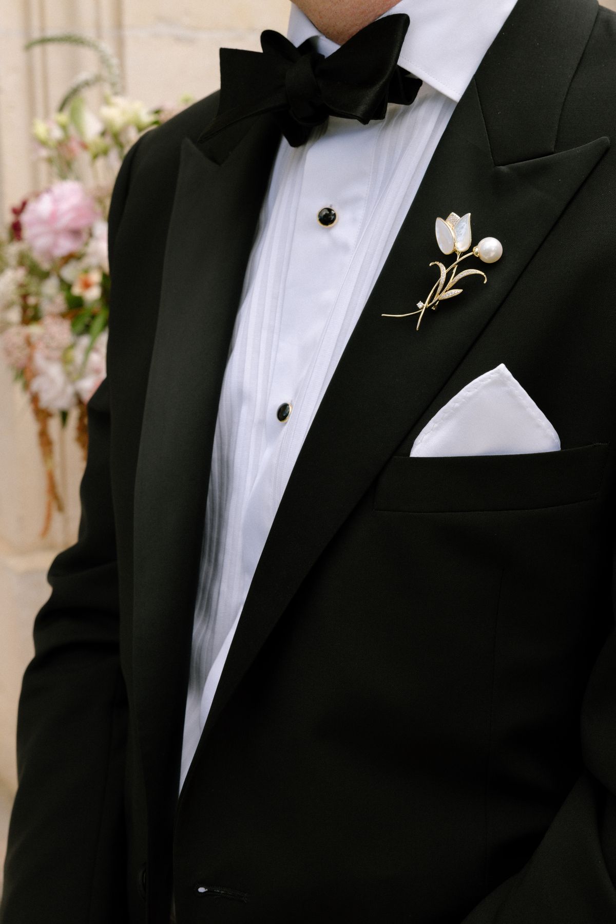 Close view of a groom's black tuxedo jacket, bow tie, white pocket square and a small white flower brooch on the lapel.