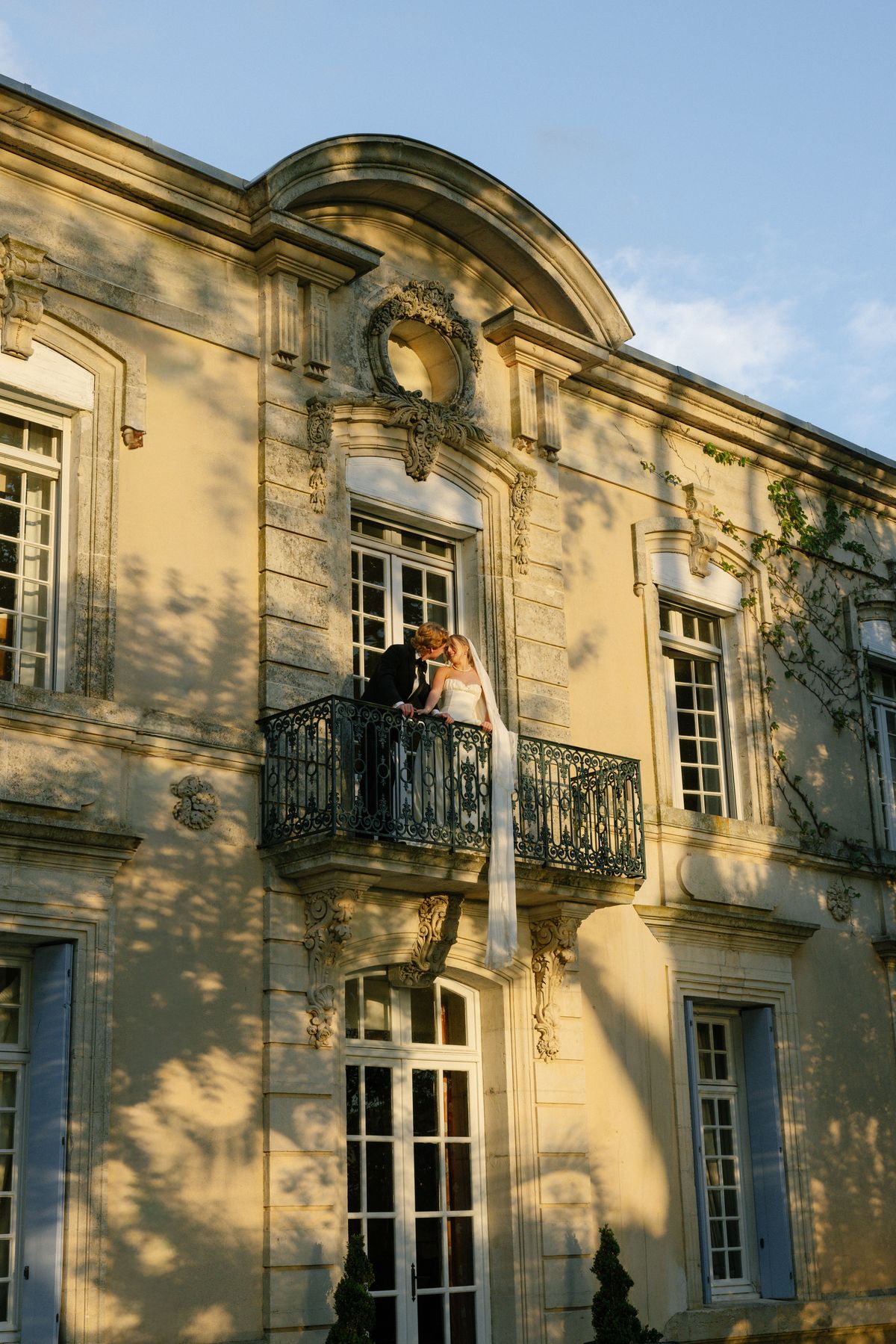 The couple kiss on a small wrought-iron balcony of a honey-coloured stone chateau under a blue sky, with a veil hanging below.