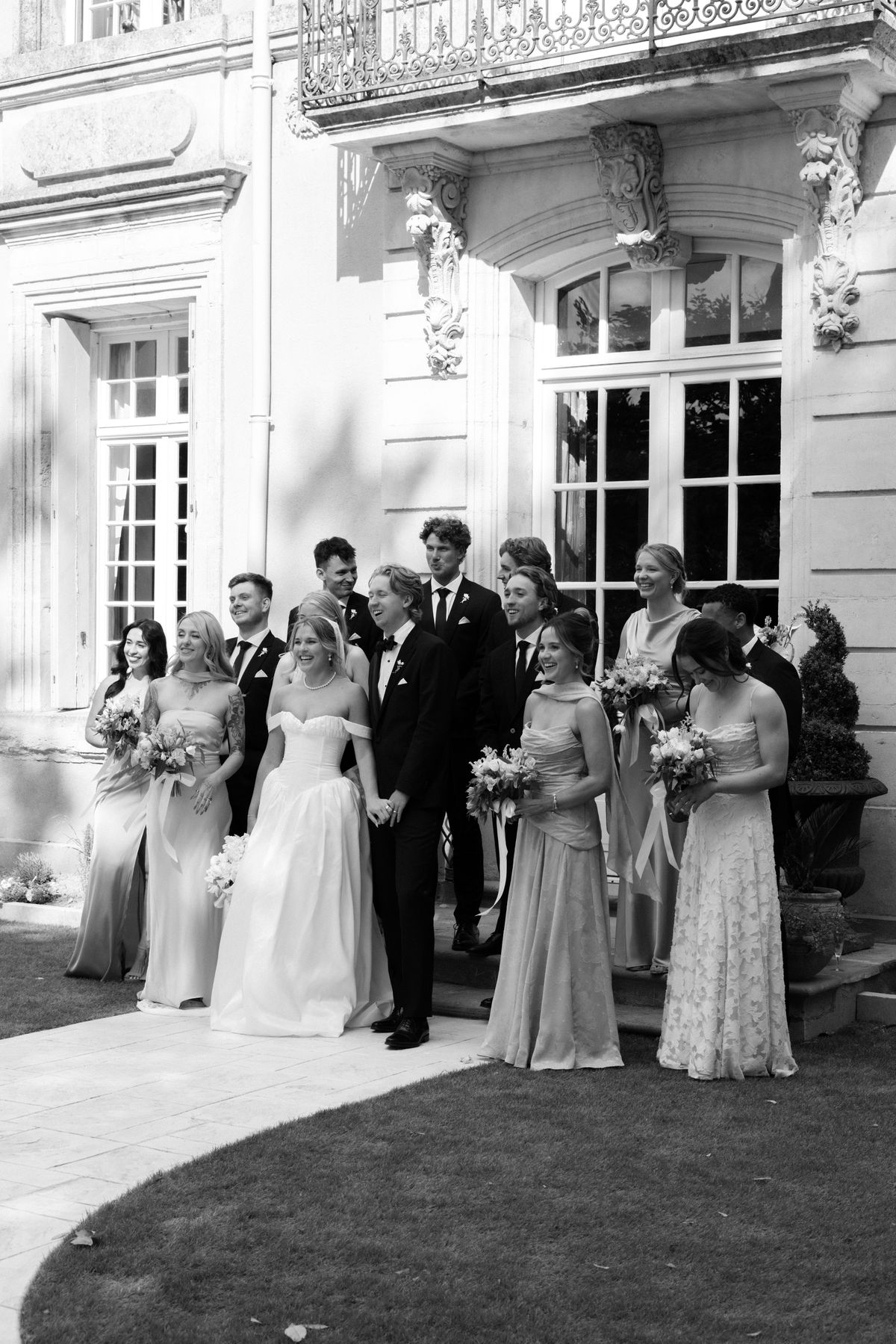 In black and white, the couple stand among bridesmaids and groomsmen in formal clothes on the steps in front of a chateau.