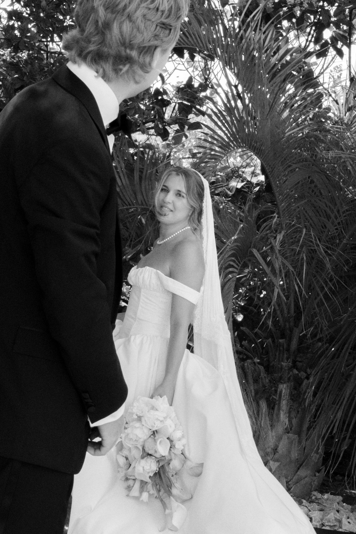 Black-and-white photo of the bride in a veil and off-shoulder gown holding roses, looking at the groom before palm leaves.