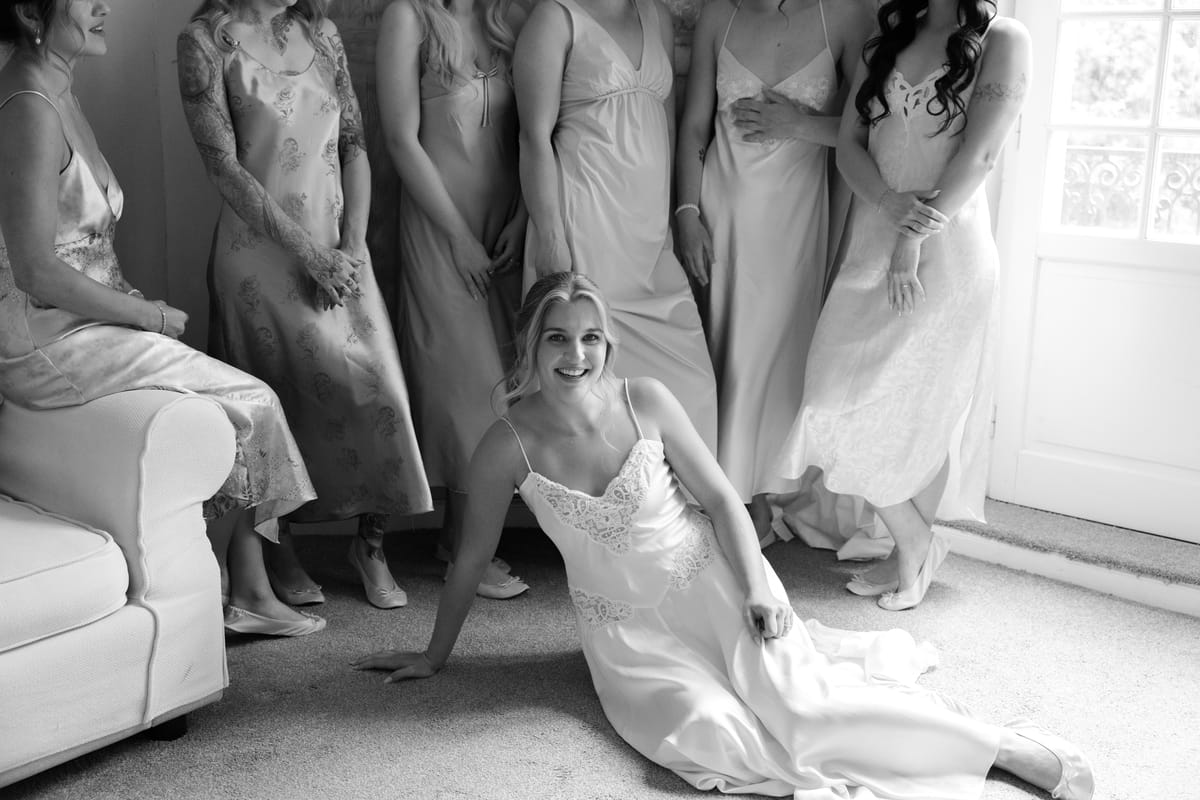The bride sits on the floor in a white slip, smiling, with her bridesmaids in pale silk dresses standing behind her.