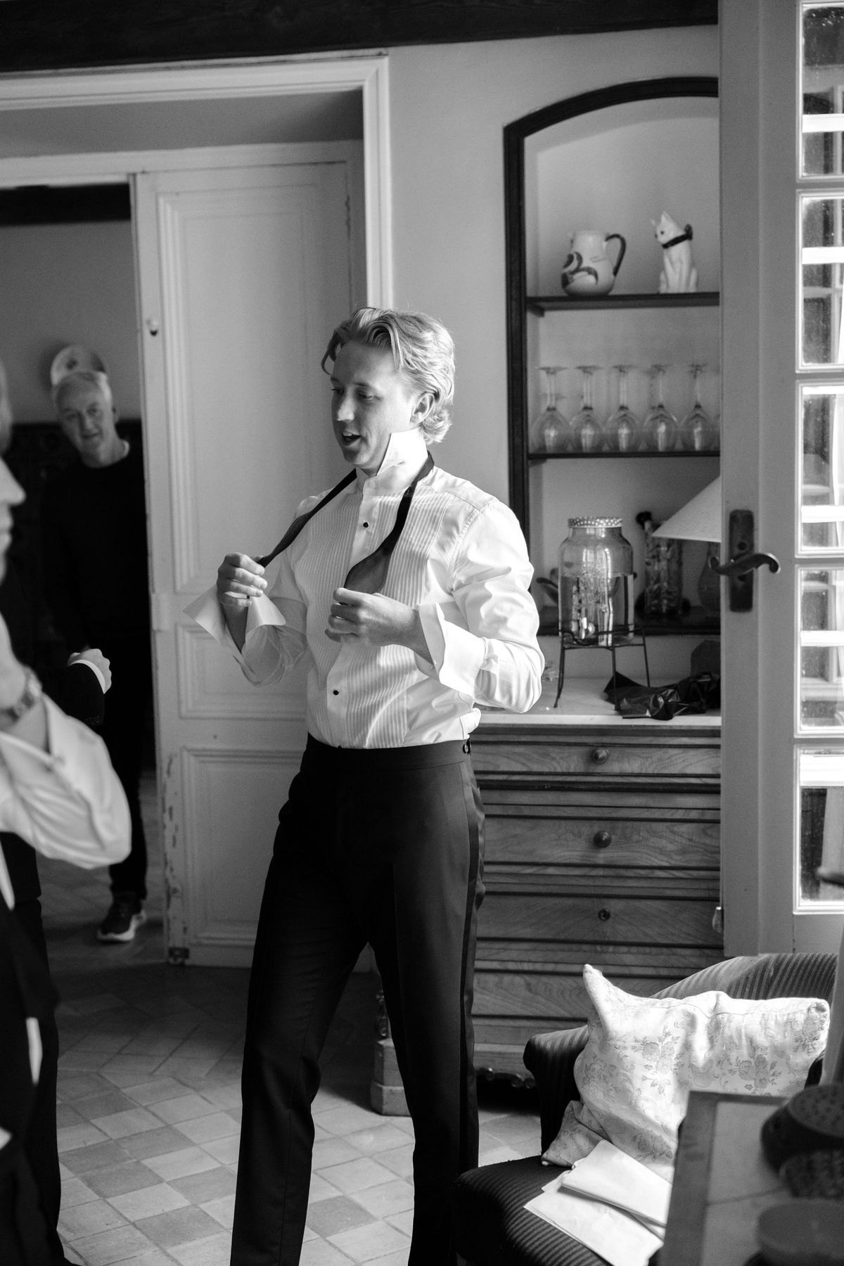 A man in a white dress shirt ties his bow tie while getting ready in a sunlit room, in black and white.
