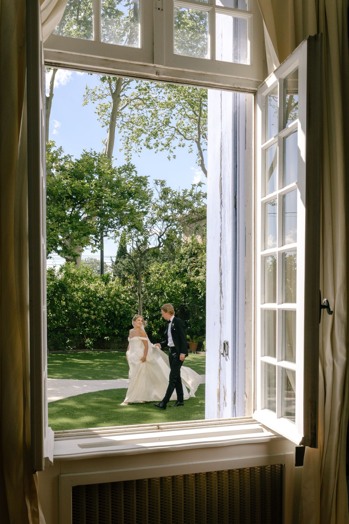 An open window with curtains frames the couple walking together across a green lawn, the bride gathering her skirt.