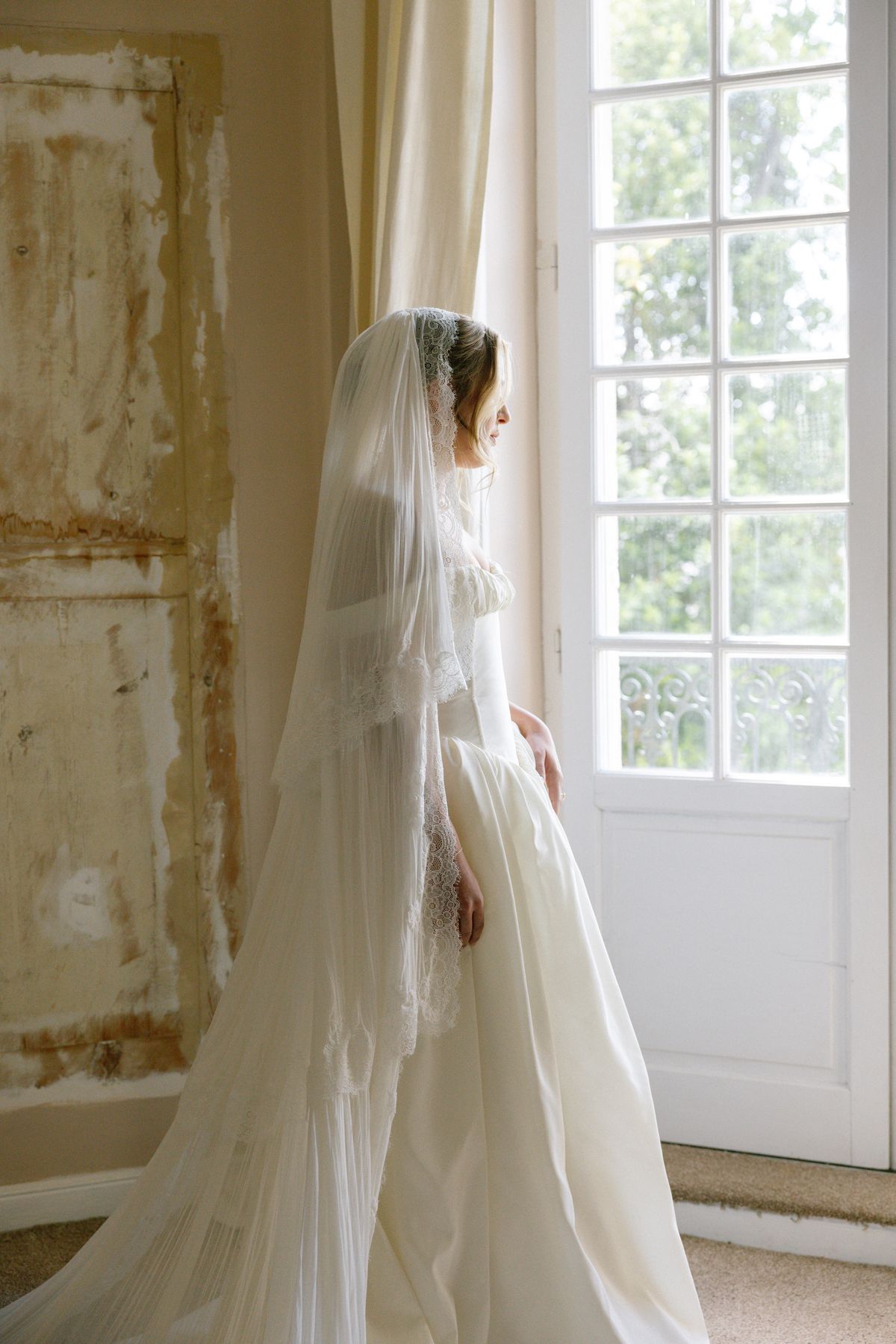 The bride, in a long lace-edged veil and ivory gown, stands in profile gazing out of a tall window beside a worn wall.