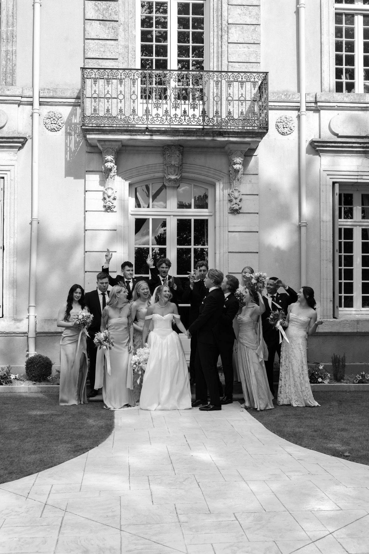 In black and white, the bride, bridesmaids and groomsmen stand together on a curved stone path in front of a grand house with a balcony.