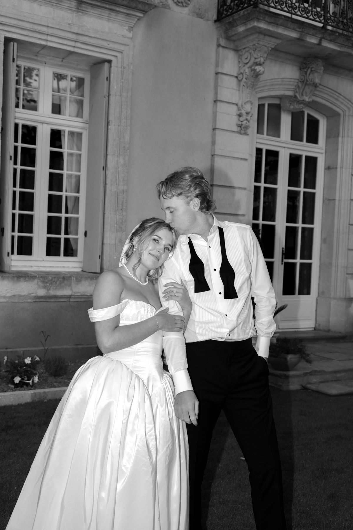 The bride in an off-the-shoulder gown leans against the groom, whose bow tie hangs open, in front of tall chateau windows.