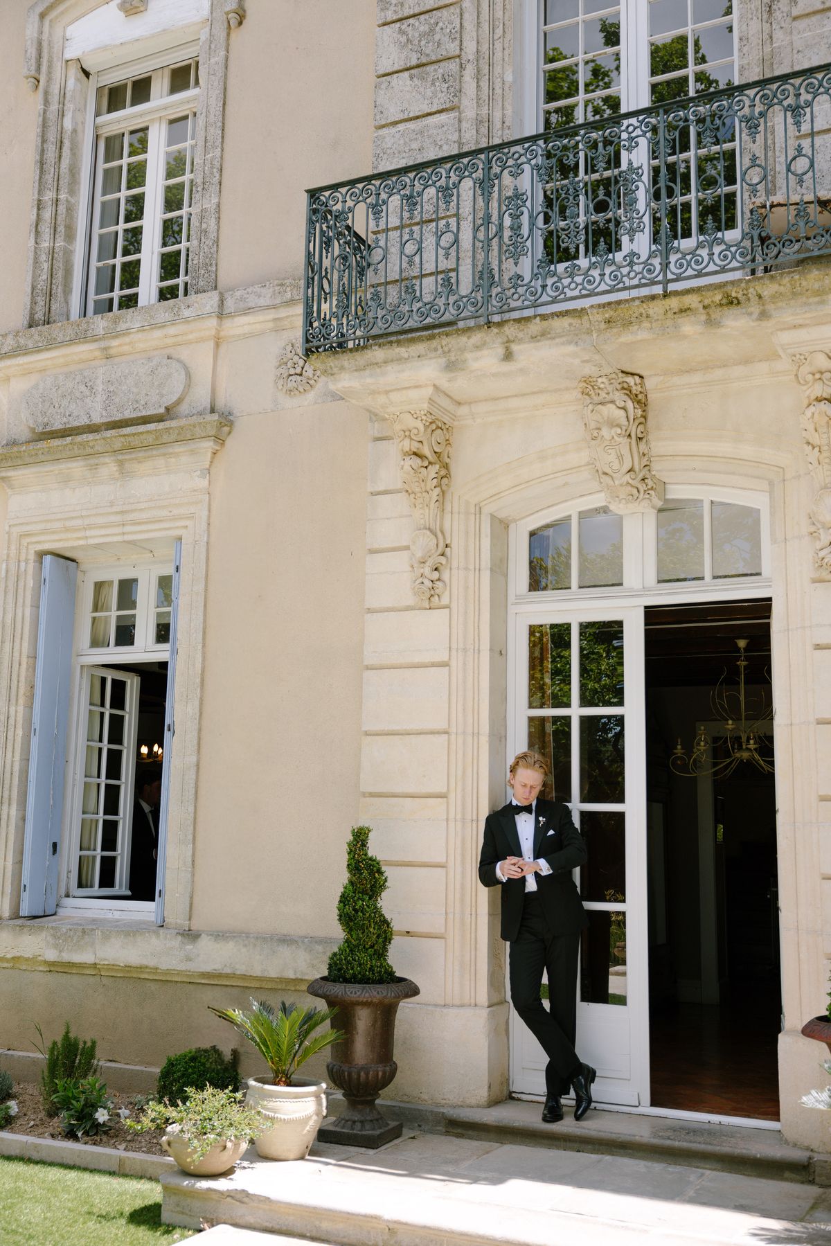 The groom in a black tuxedo leans casually against the frame of an open French window on a pale stone chateau facade.